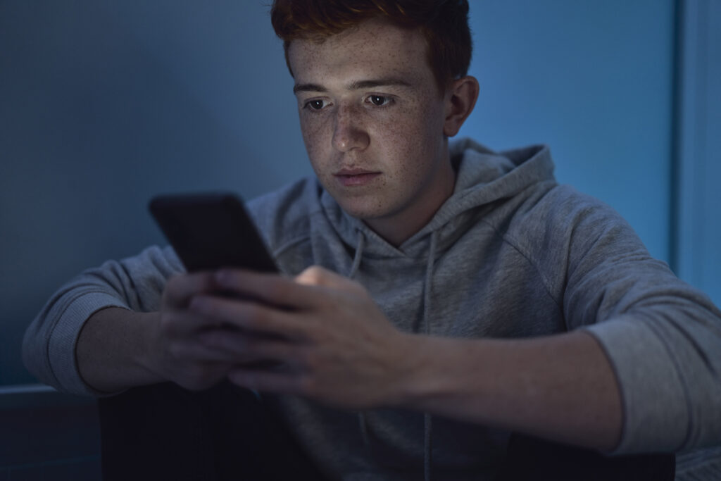focus caucasian teenage boy using mobile phone while sitting at night in his room