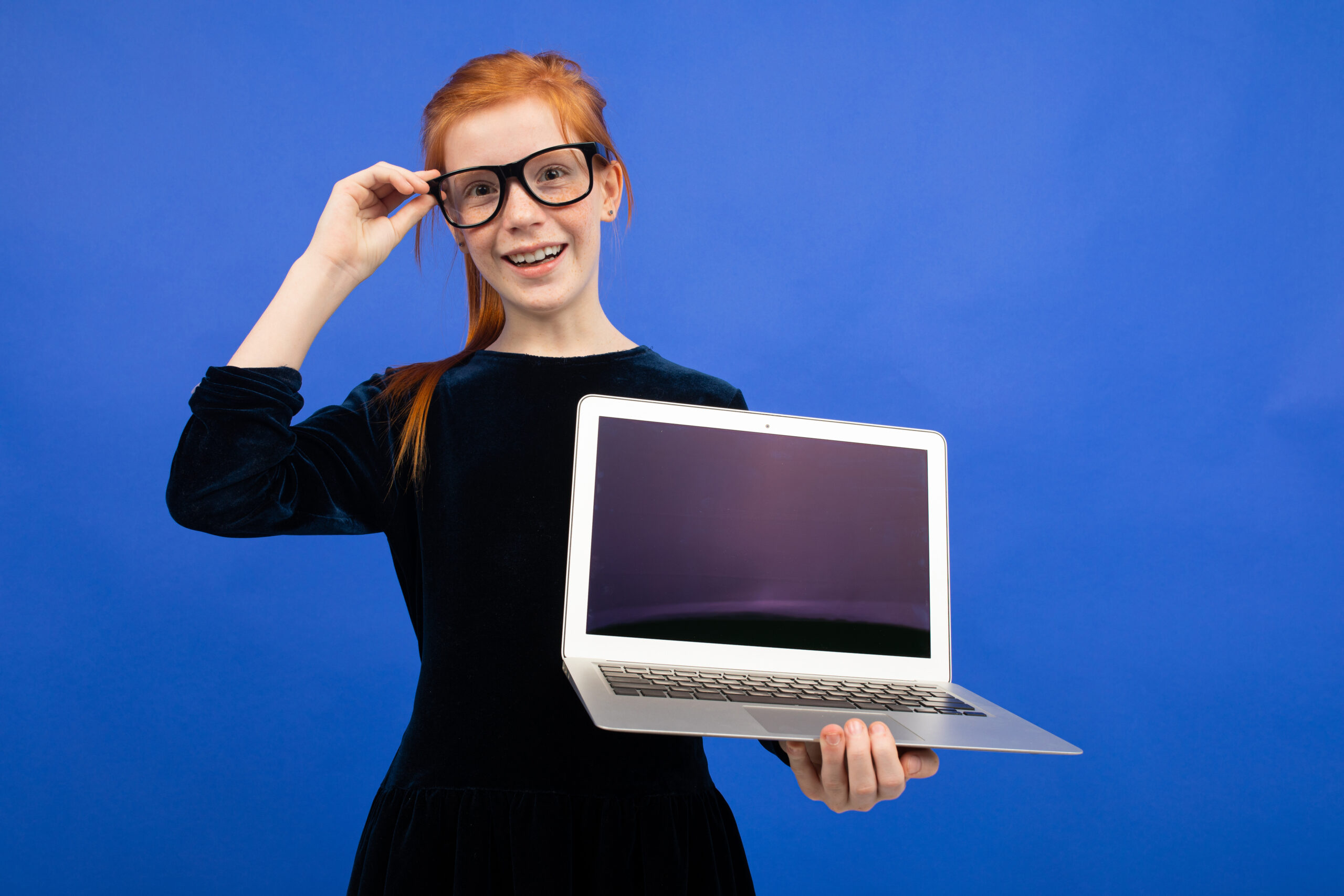 Home smart red haired teenager girl in glasses holds a laptop with a blank screen mock up to insert a page on a blue background